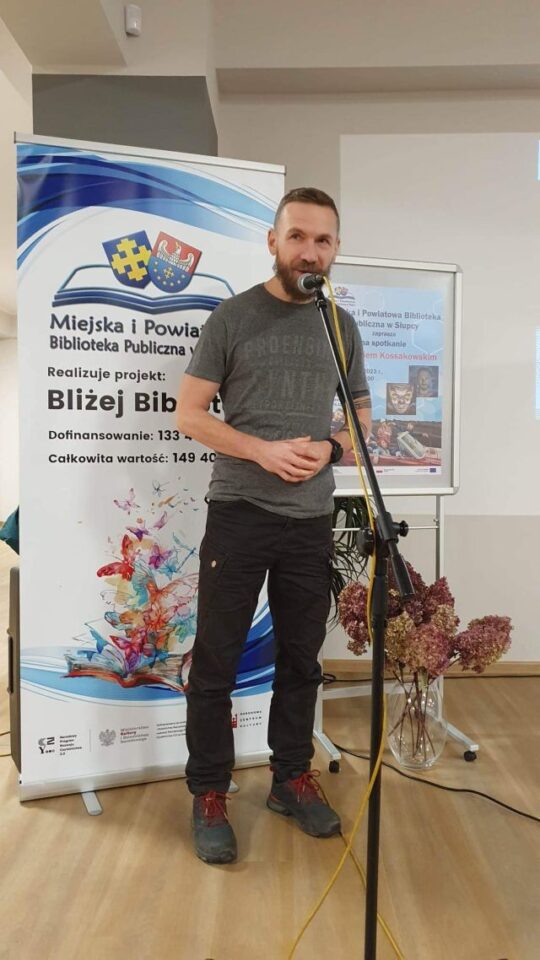 mężczyzna w siwym podkoszulku i czarnych spodniach stoi przed planszą z nazwą projektu