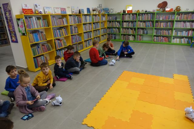na środkul leżą duże pomarańczowe puzle, wokół nich na podłodze siedzą dzieci, a przed nimi białe małe roboty, w tle regały z książkami