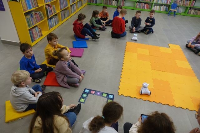 na środku leżą duże pomarańczowe puzle, wokół nich na podłodze siedzą dzieci, a przed nimi białe małe roboty, w tle regały z książkami