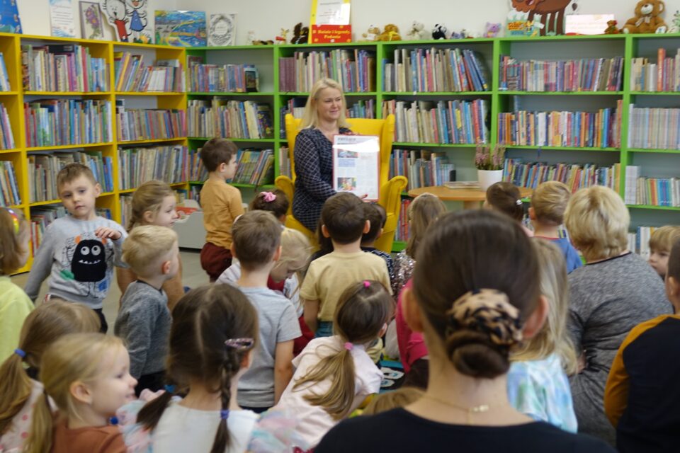dzieci siedzą na dywanie, przed nimi kobieta w siwej sukience siedzi na żółtym fotelu, w tle regały z książkami