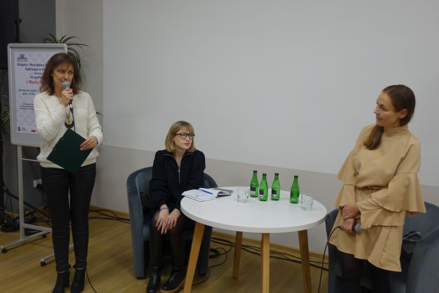 diwe kobiety stoją jedna w białym swetrze trzyma mikrofon druga w kremowej sukience słucha, kobieta w czarnej sukience siedzi przy białym stoliku okrągłym