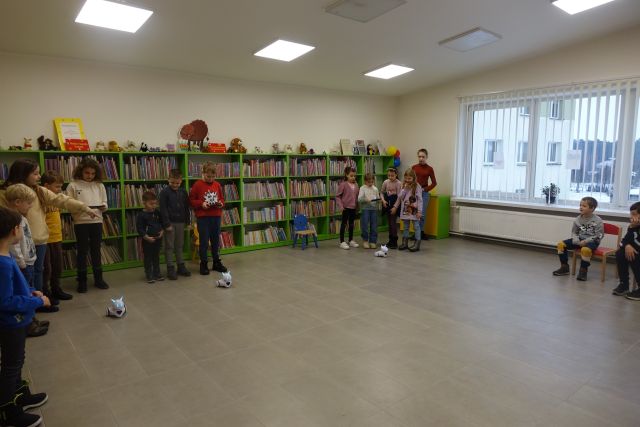 dzieci stoją i za pomocą tabletów sterują robotami, które jeżdżą po podłodze, w tle regały z książkami