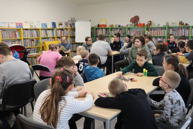 około 30 dzieci siedzi przy stolikach i słucha jak mężczyzna siedzący na żółtym fotelu czyta książkę, w tle regały z książkami