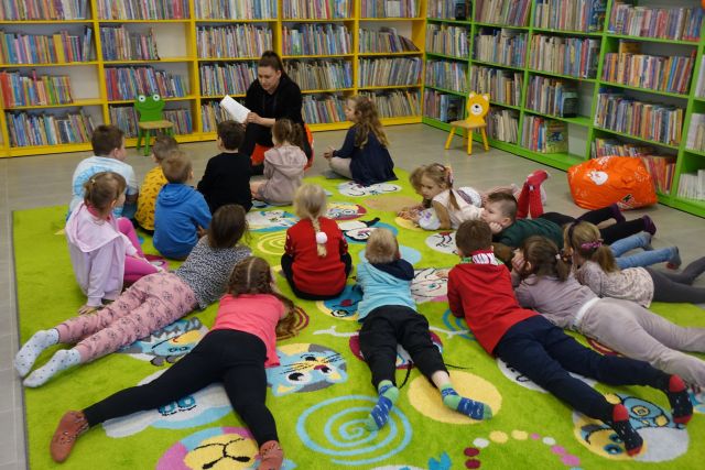 dzieci leżą na zielonym dywanie i słuchają czytaną im książkę przez kobietę w czarnym swetrze, w tle regały z książkami