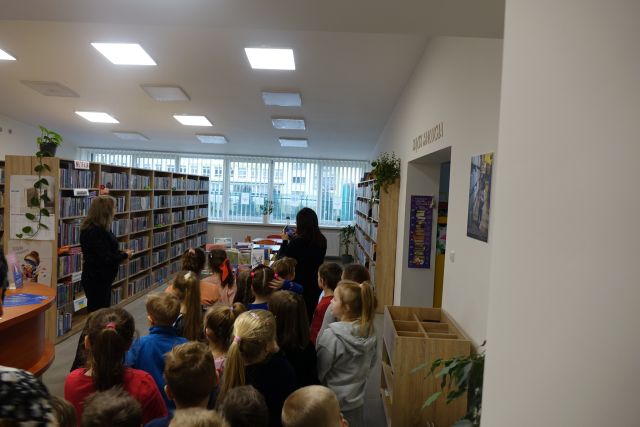 dzieci w sali bibliotecznej, przed nimi stoliki i krzesła oraz regały z książkami