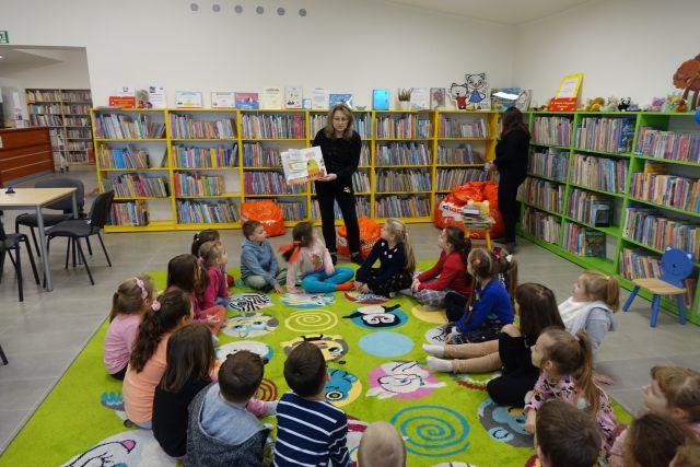 dzieci siedzą na zielonym dywanie, przed nimi stoi kobieta i pokazuje książkę, w tle regały z książkami