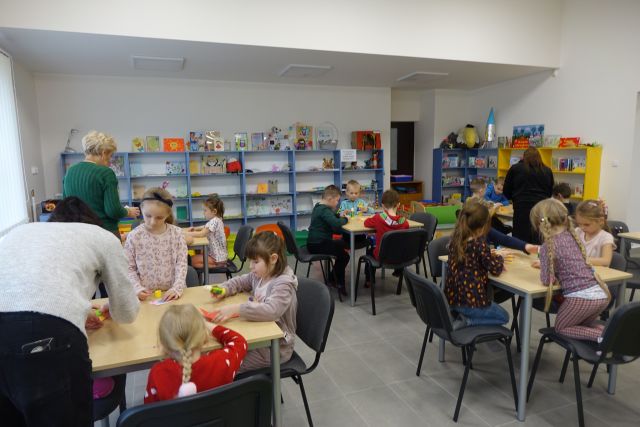 dzieci siedzą przy stolikach i wykonują kolorowe etui na kartę biblioteczną, w tle kolorowe regały