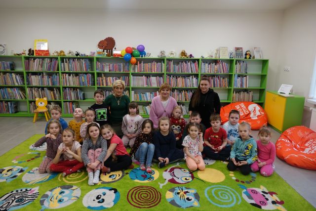 dzieci siedzą na zielonym dywanie i pozują do zdjęcia grupowego z 3 opiekunkami, w tle regały z książkami