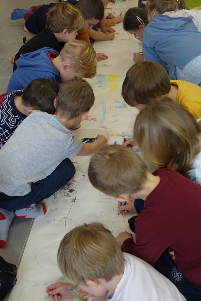 dzieci na długim papierze położonym na podłodze malują kredkami