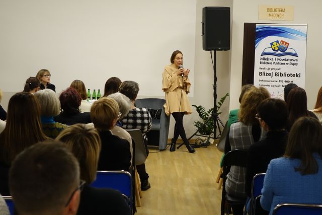 uczestnicy spotkania siedzą na krzesłach, przed nimi kobita w beżowej sukience stoi z mikrofonem