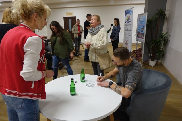 mżczyzna siedzi przy stole i wpisuje dedykację uczestnicce spotkania, w tle inni uczestnicy