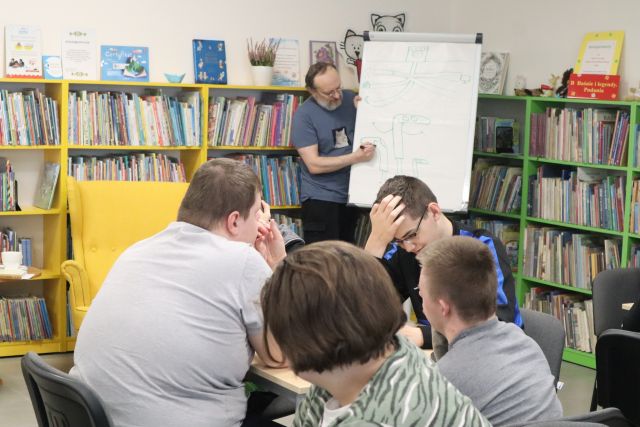 mężczyzna na flipcharcie rysuje, przy stolikach siedzą dzieci i rysują, w tle regały z książkami