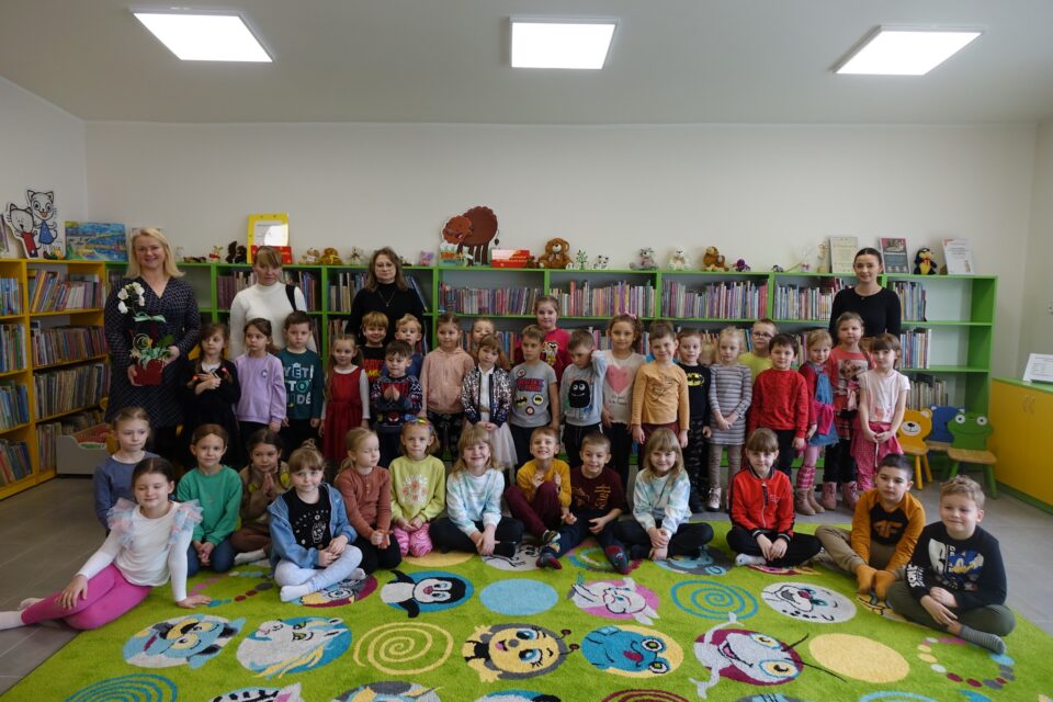 dzieci i kobiety uczestniczące w spotkaniu pozują do zdjęcia grupowego, w tle regały z książkami