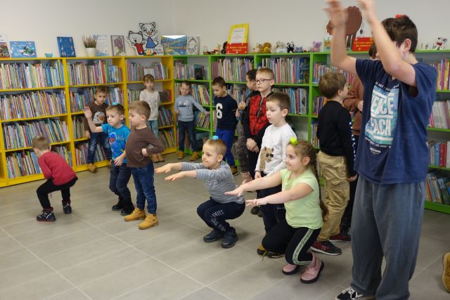 dzieci z klockiem na kłowie wykonuja przysiady, w tle regały z książkami