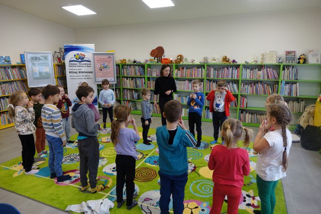dzieci i kobieta w czarnej bluzce i czarnych spodniach stoją na zielonym dywanie, w tle regały z książkami