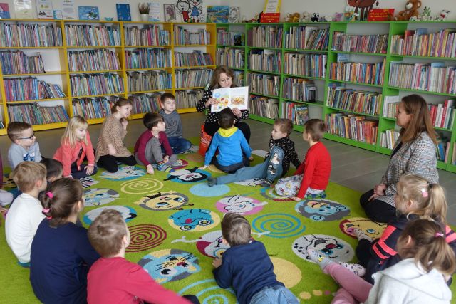 dzieci siedzą na dywanie, przed nimi siedzi kobieta i pokazuje książkę, w tle regały z książkami