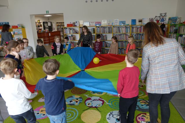 dzieci i dwie osoby dorosłę trzymają dużą kolorową chustę, na której leży żółty balon, w tle regały z książkami