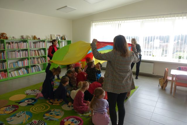trzy kobiety unoszą do góry kolorową dużą chustę, pod nią siedzą dzieci na dywanie, w tle regały z książkami