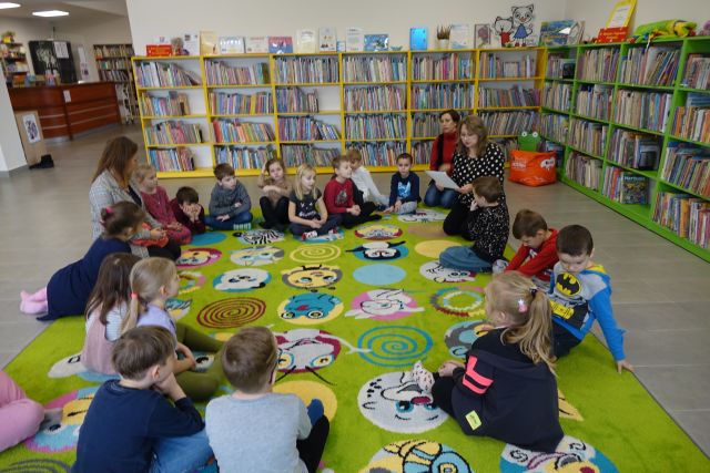 dzieci siedzą na zielonym dywanie, pomiędzy nimi 3 kobiety, w tle regały z książkami