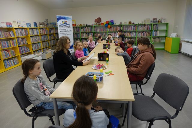 dzieci i osoba dorosła siedzą przy w rzędzie ustawionych stołach, na stołach mają kredki, w tle regały z książkami