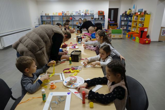 dzieci siedzące przy stołąch wykonują ramki na zdjęcia, pomagają im osoby dorosłe