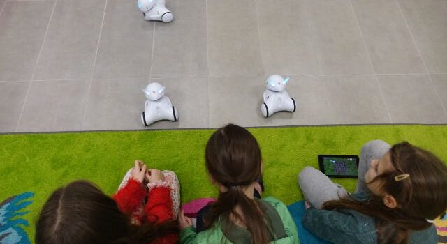 dziewczynki siedzą na zielonym dywanie, trzymają w dłoniach tablety i sterują nimi robotami, któe są na podłodze