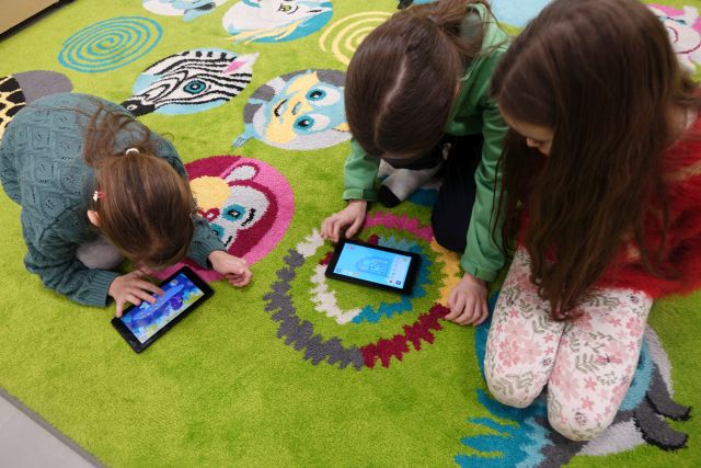 3 dziewczynki siedzą na zielonym dywanie i grają na tabletach
