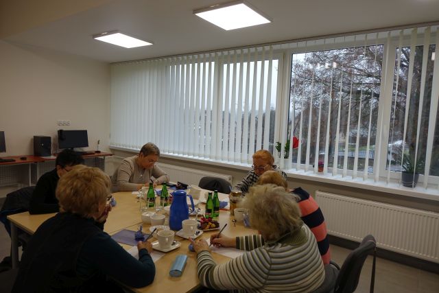 seniorki siedzą przy stołach i rozwiązują krzyżówki, w tle biurka z komputerami