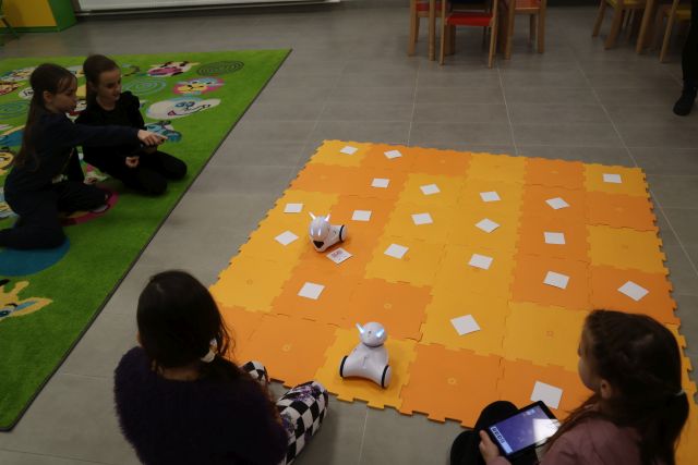 dzieci siedzą wokół żółtej maty, pok któej jeżdżą małę białe roboty i leżą kartki