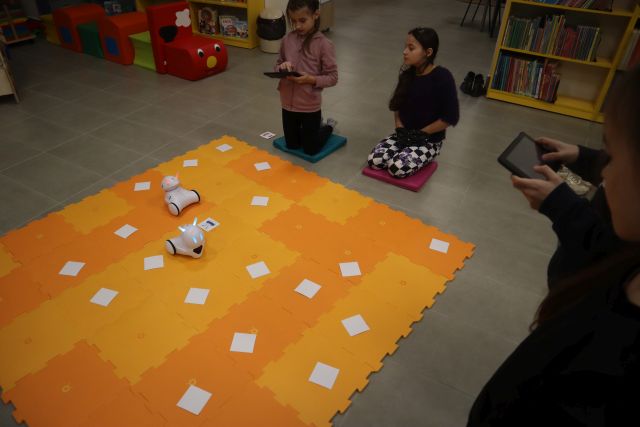 dzieci siedzą wokół żółtej maty, pok któej jeżdżą małę białe roboty i leżą kartki