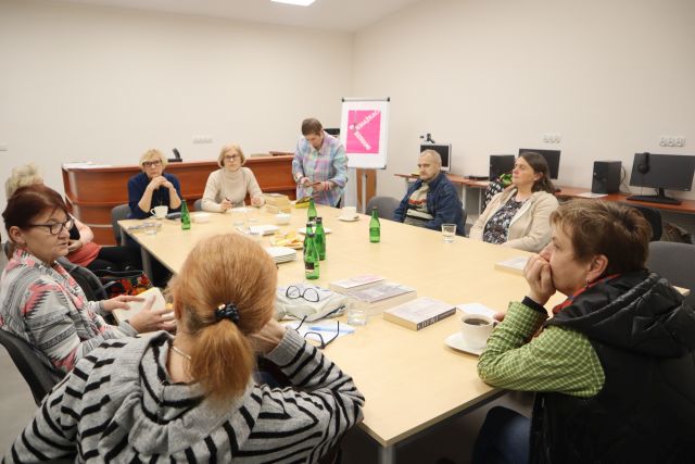uczestnicy spotkania, 9 osób siedzi przy stołach połączonych, w tle biurka z komputerami