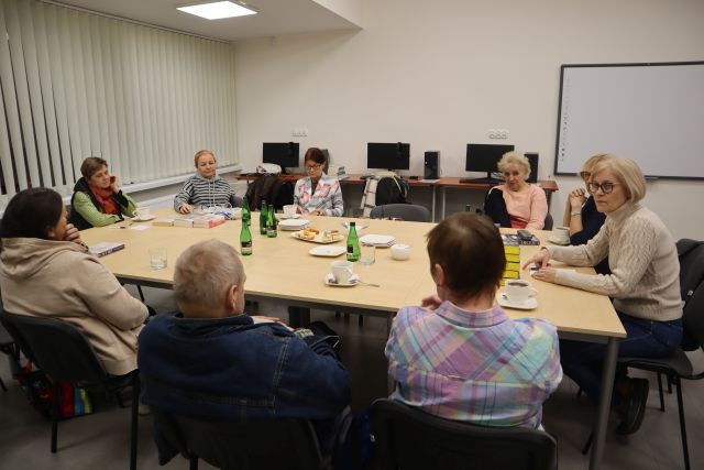 uczestnicy spotkania, 9 osób siedzi przy stołach połączonych, w tle biurka z komputerami