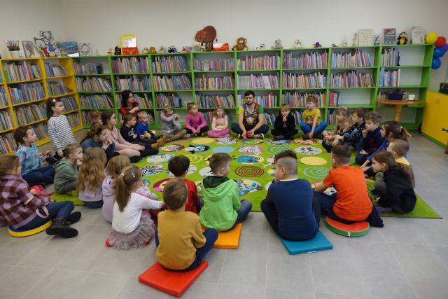 dzieci i mężczyzna siedzą w okręgu na dywanie zielonym, w tle regały z książkami