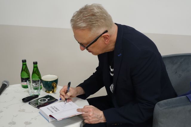 mężczyzna w granatowej marynarce i okularach podpisuje książkę