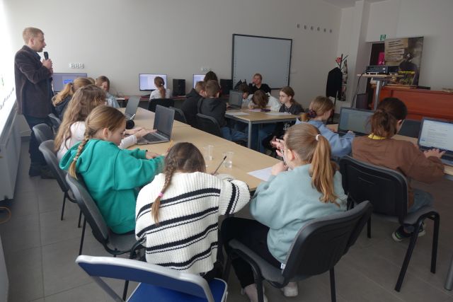 młodzi uczestnicy warsztatów siedzą przy stołąch i piszą, przed nimi stoją laptopy i komputery