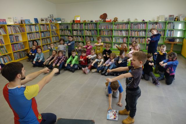 dzieci siedzą na zielonym dywanie z wyciągniętymi rękami, w tle regały z książkami