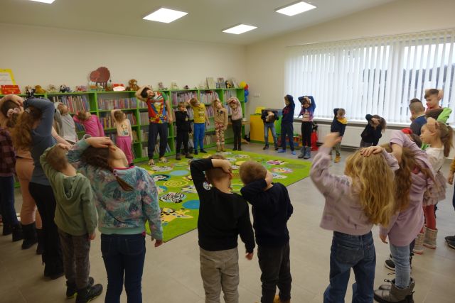 dzieci stoją w okręgu i wykonują ćwiczemia, w tle regały z książkami