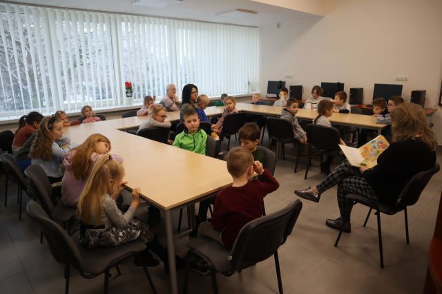 uczestnicy warsztatów, 28 dzieci siedzą przy stołach i suchają czytającej bajkę kobiety siedzącej przed nimi na krześle 9 w tle biurka z komputerami