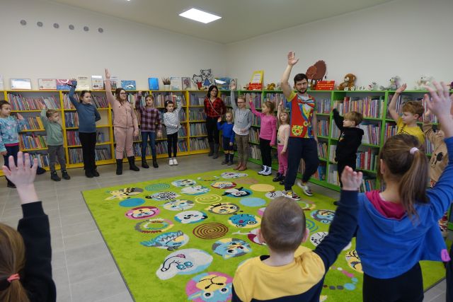 dzieci stoją w okręgu i wykonują ćwiczemia, w tle regały z książkami