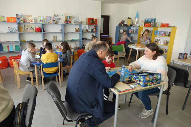 dzieci i osoby dorosłe grają w gry planszowe, w tle regały