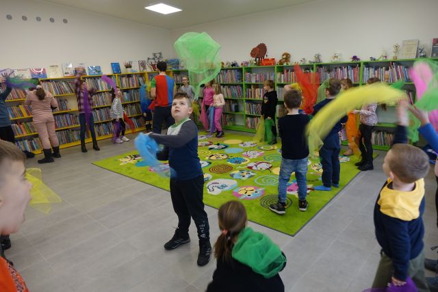 dzieci podrzucają do góry kolorowe chusty, w tle regały z książkami