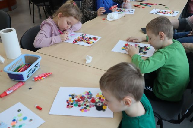 dzieci siedzą przy stołąch i wyklejają na kartkach guzikami misie