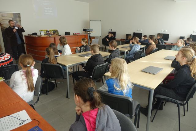 młodzi uczestnicy warsztatów siedzą przy stołach i słuchają mężczyzny prowadzącego warsztaty. w tle biurka z komputerami