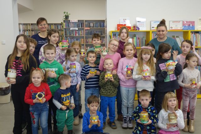 dzieci uczestniczące w warsztatach i dwie osoby dorosłe pozuja do zdjęcia z wykonanymi przez siebie lampionami ze sznurka słoików i różnych ozdób, w tle regały z książkami