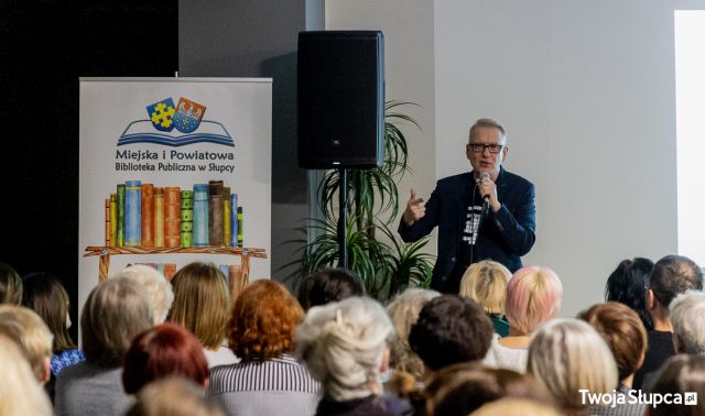 uczestnicy spotkania siedzący tyłem do aparatu, przed nimi autor z mikrofonem, z lewej strony głośnik i plansza z logo biblioteki