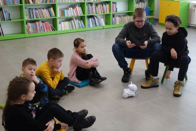 dzieci siedzą na podłodze i na krzesełka, chłopiec za pomoca tabletu steruje robotem, w tle regały z książkami
