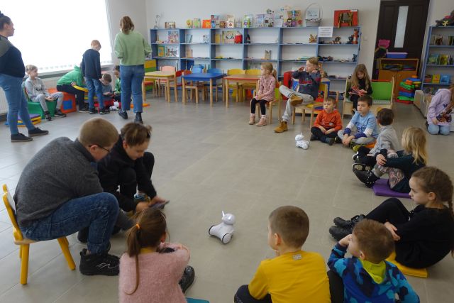 dzieci siedzą na podłodze, pomiędzy nimi jeżdżą małe białe roboty, w tle regały z książkami