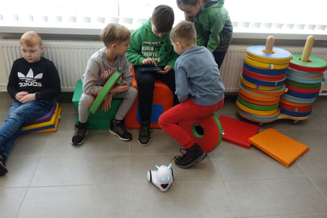 dzieci siedzą na kolorowych siedziskach i sterują za pomocą tabletu robotem