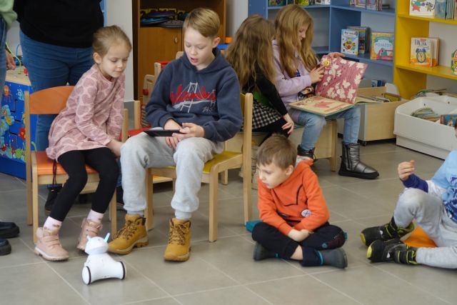 dzieci siedzą na podłodze i krzesłach i sterują za pomocą tabletu robotami