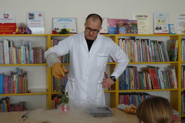mężczyzna w biały fartuchu i żółtej rękawicy macza czerwoną różę w azocie, w tle regały z książkami